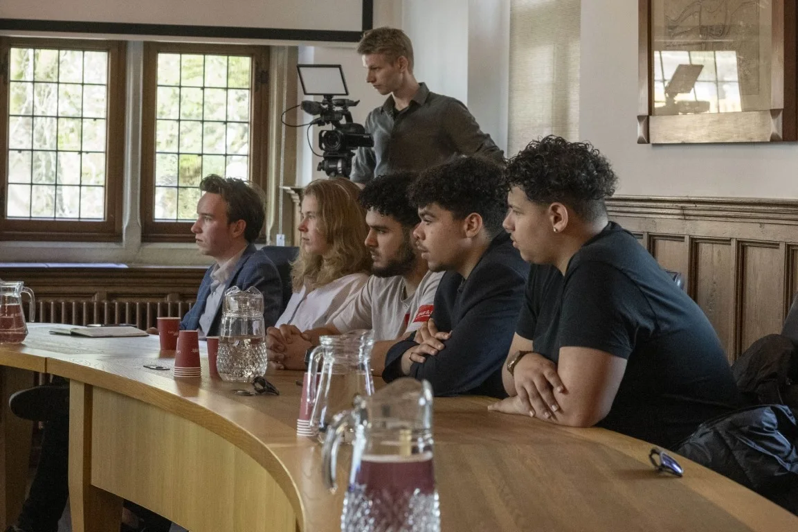 Vijf jonge mensen luisteren aan de gebogen tafel in een raadzaal, achter hen staat een cameraman
