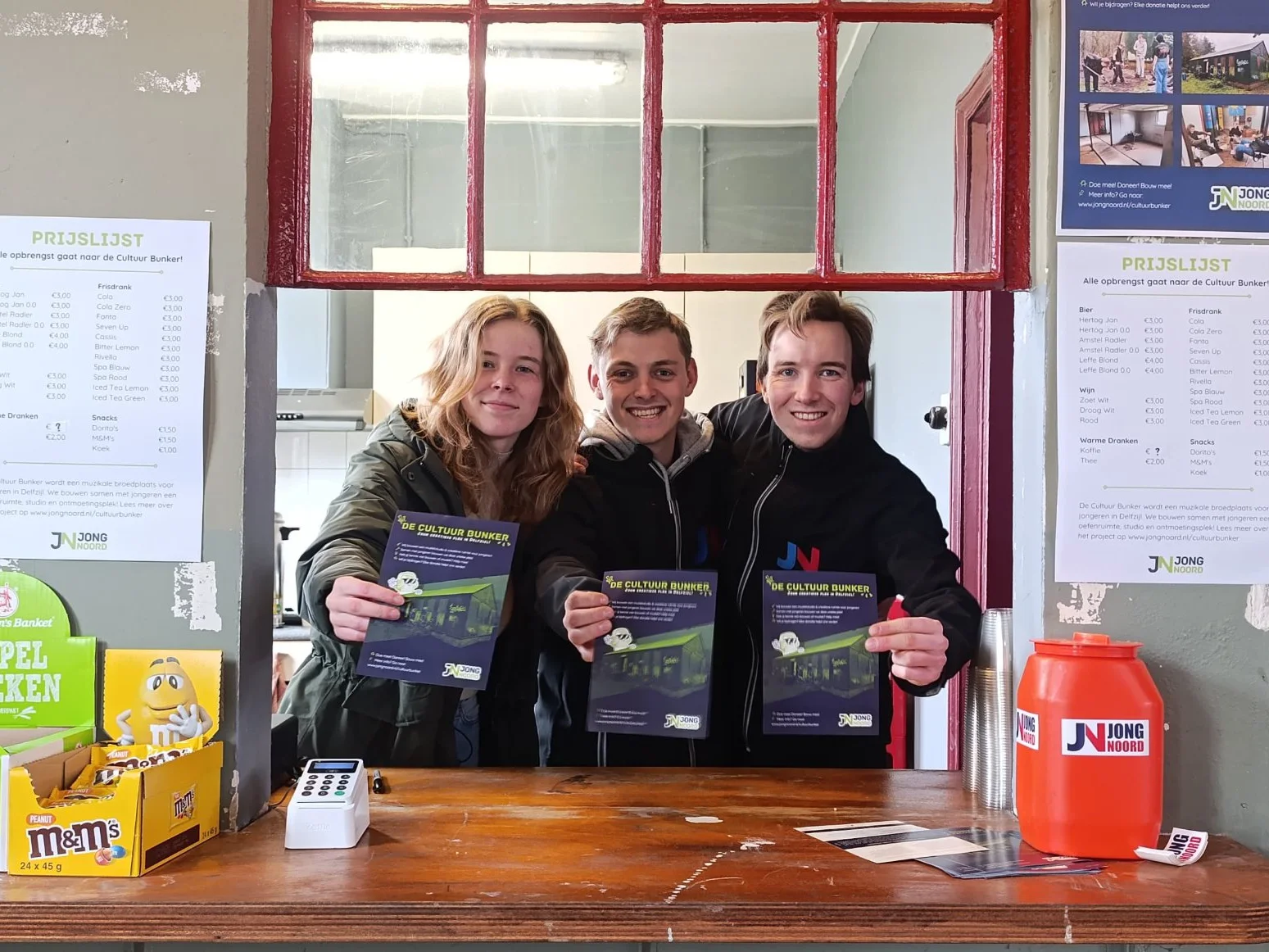 Drie jonge mensen achter de bar met flyers van de Cultuur Bunker. Op de prijslijst staat dat alle opbrengst naar de Cultuur Bunker gaat.